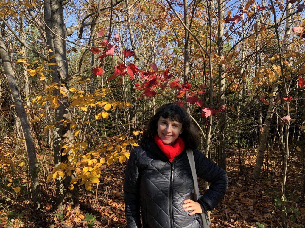 A woman with black, shoulder-length hair, wearing a red wool scarf tucked in the neck of a black puffy jacket, smiles, left hand on her waist. She is surrounded by yellow and green leaves of trees in the woods around her.