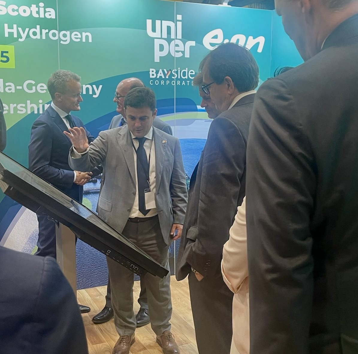 EverWind CEO and founder, Trent Vichie, in a grey suit, dark blue tie and white shirt, stands with his right hand raised, in the centre of this photo appears to be explaining a display board to other men at the occasion of the signing of memoranda of understanding with Uniper and E.On.
