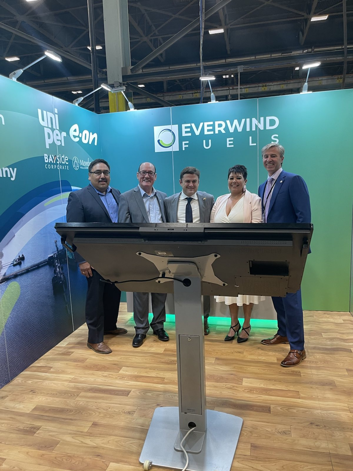 Four men and one woman lined up at a podium, all smiling. Premier Tim Houston (right), EverWind CEO and founder Trent Vichie (centre), Membertou First Nation Chief Terrance Paul (2nd from left), and other dignitaries at the signing of the MOU with Uniper and E.On.