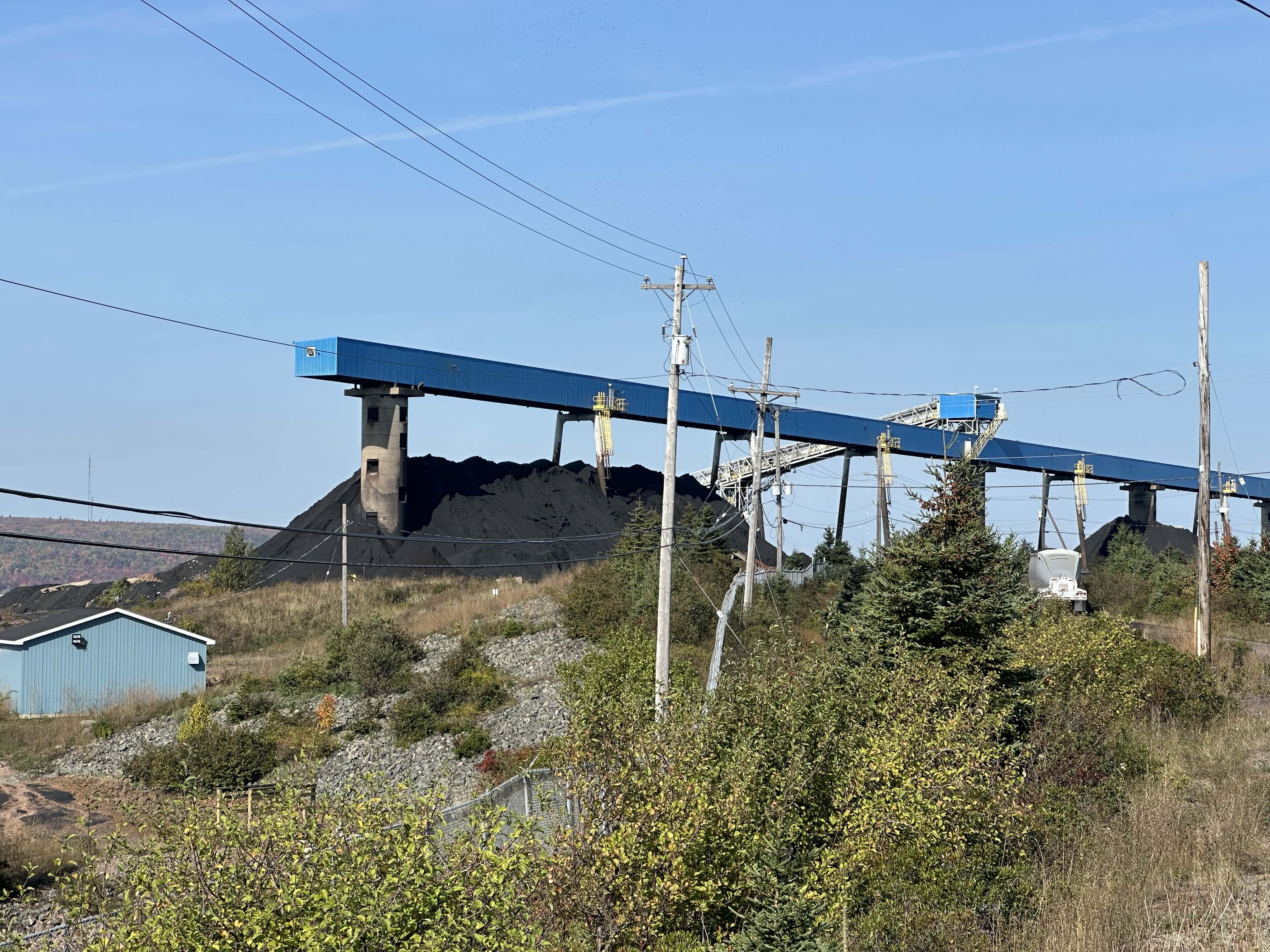 Under a blue sky, and surrounded by power lines, is a very large pile of black coal. The area is enclosed by a chain link fence, and scrubby pale green vegetation. Over the pile of coal is a mounted blue tunnel structure, with what looks like a conveyor leading up to it.