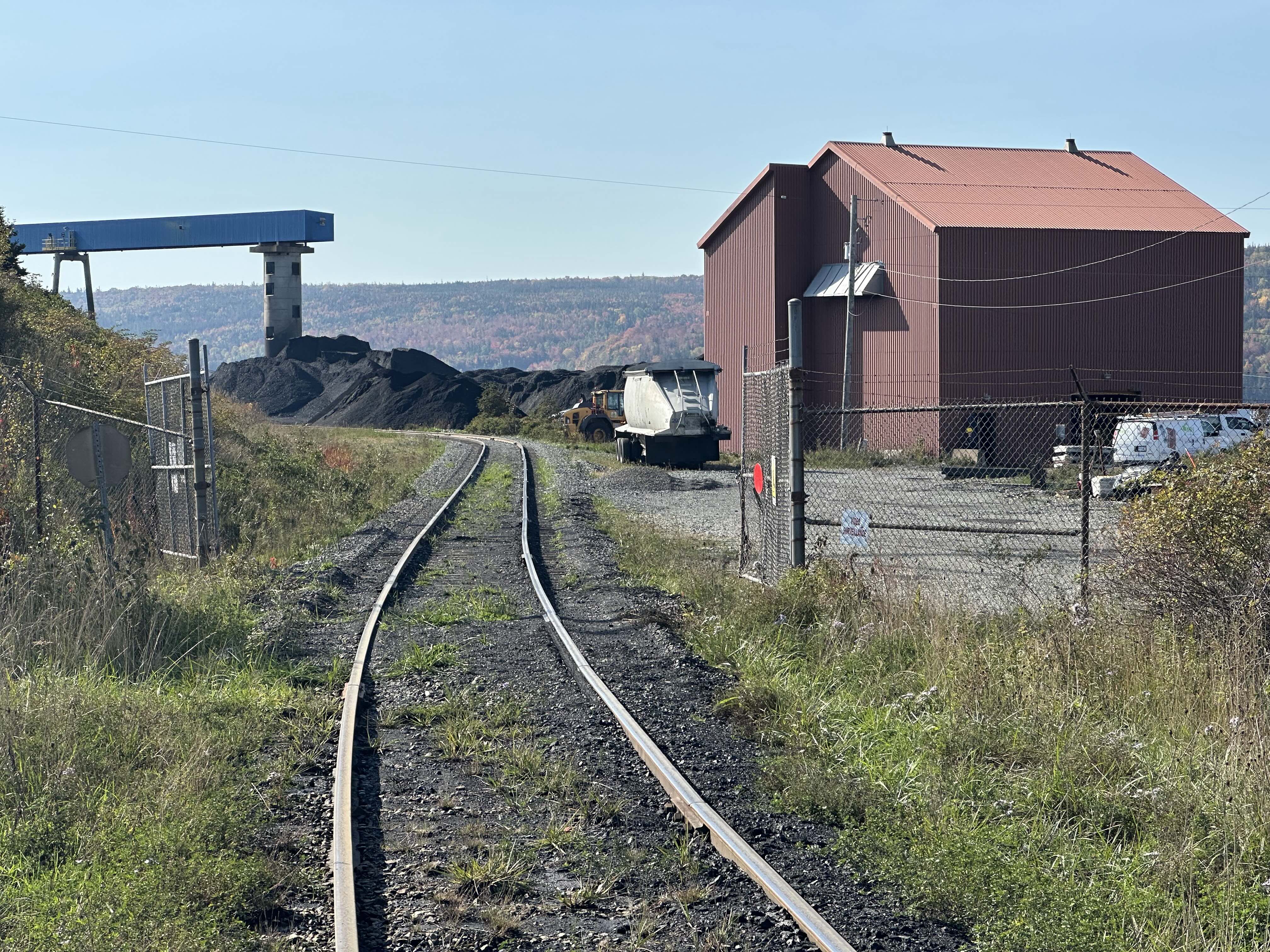Sighting the camera down railroad tracks, which lead towards and then veer left of a very large black heap of coal. The tracks lead through an open gate in a chain link fence. On the right is a blocky windowless brown building, with a brick-coloured roof, with a white tanker trailer parked beside it. On the left is a tower, suspending a blue enclosed tunnel.