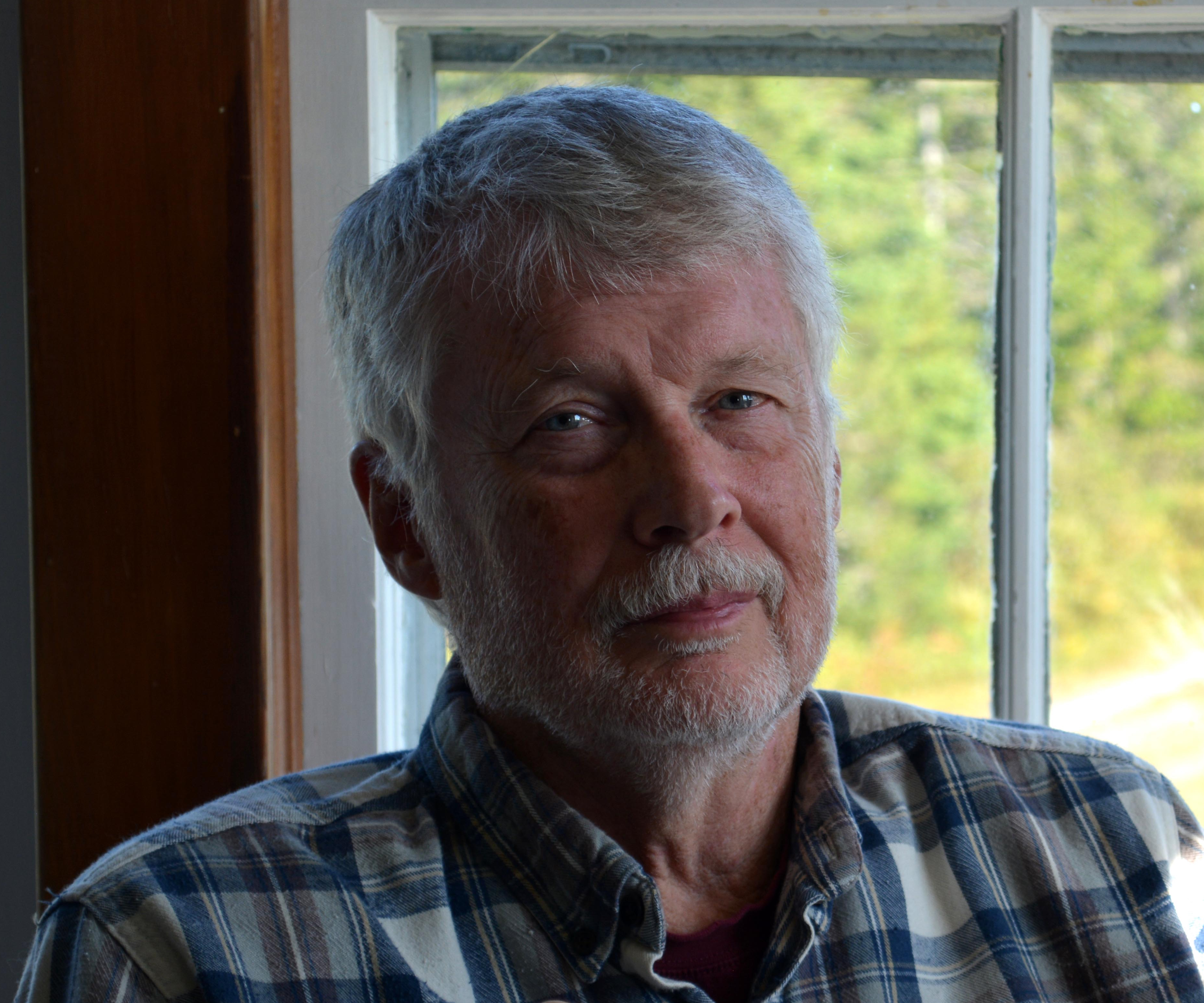 A portrait of a man with short grey hair, mustache and light beard, wearing a plaid shirt, looks intently at the camera, with his mouth closed. Behind him, out of focus, is greenerg through a white frame window.