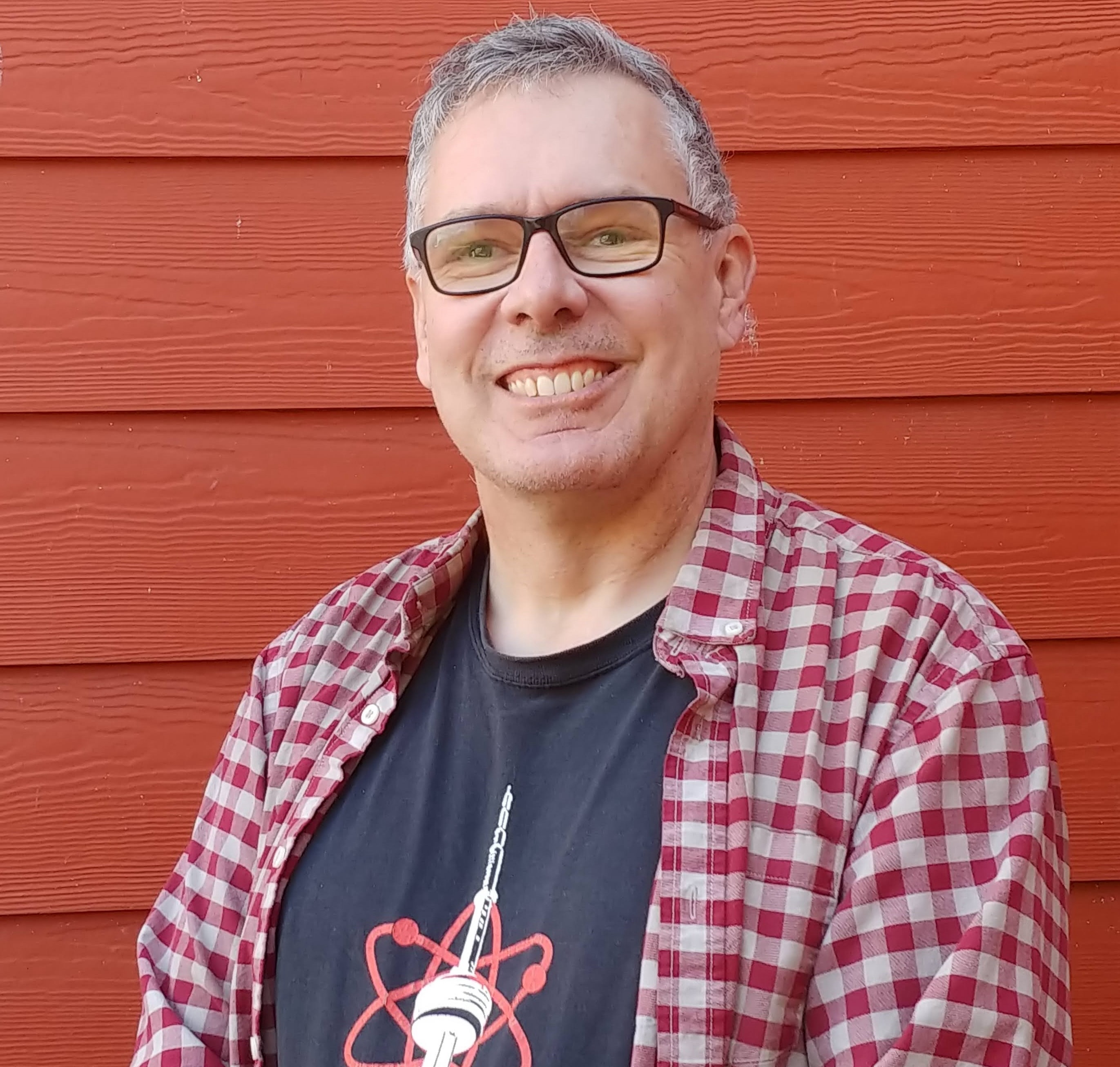 A smiling man with short greyish hair, glasses, and wearing a white and red chequered shirt open to show a black t-shirt with atoms revolving around Toronto's CN Tower and the words "for science, Toronto" showing.