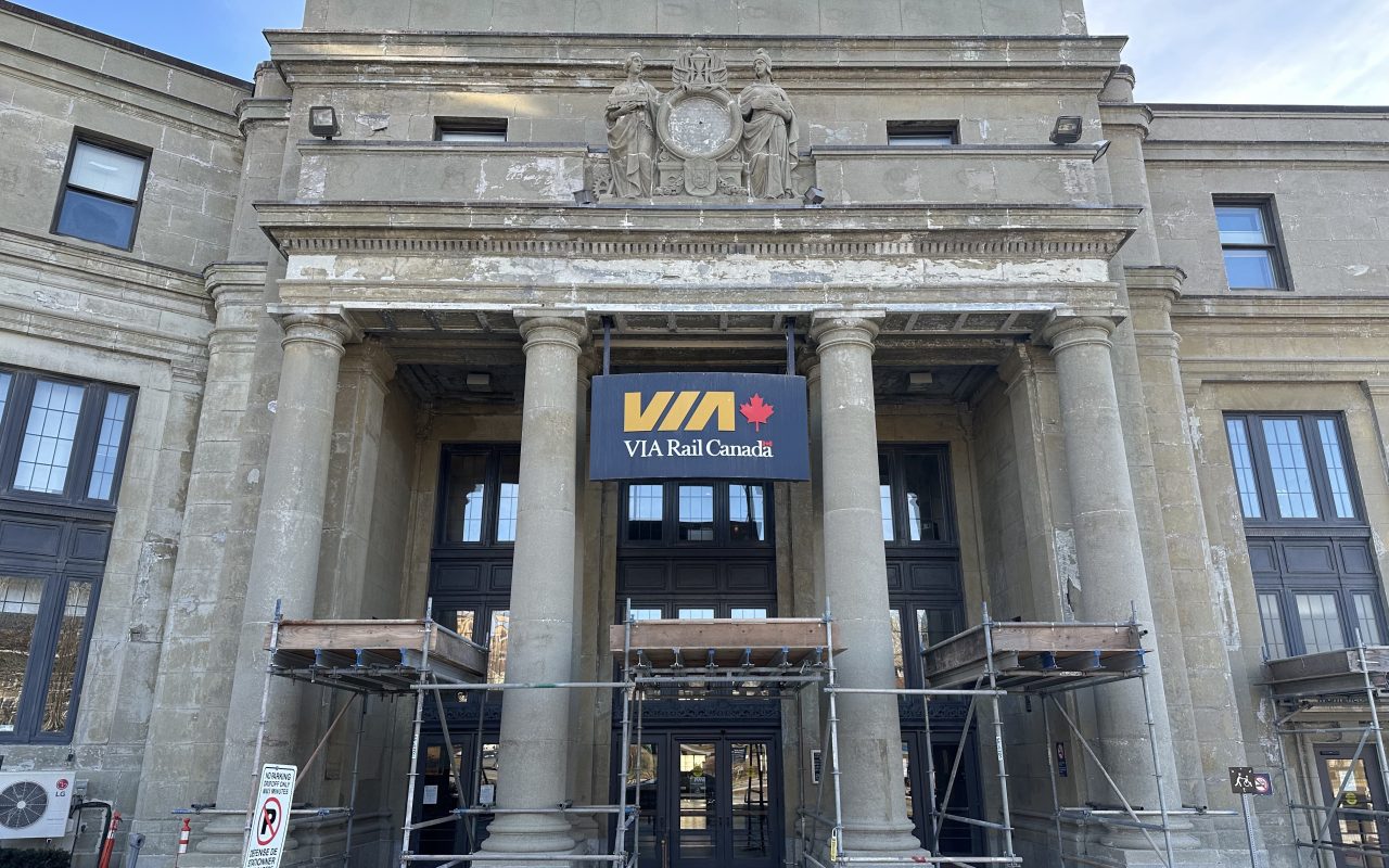 Grandiose facade of a concrete building, with pillars and empty place where a clock may once have been. The sign "VIA Rail Canada" with the yellow logo and red maple leaf is suspended between two columns, and construction is ongoing, as scaffolding is in place between the columns. Credit: Joan Baxter