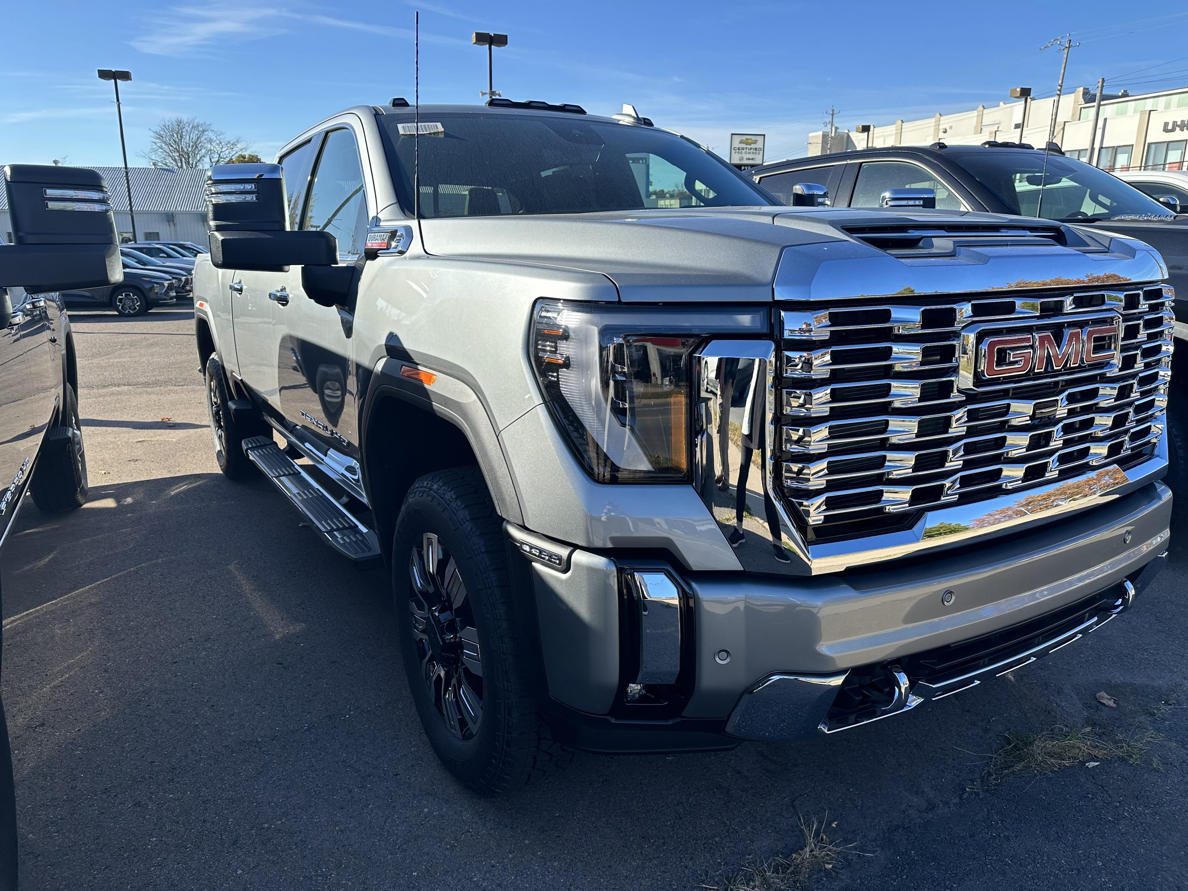 A new and very shiny large silver-grey pickup in a car dealer lot, with a massive chrome grille, and the letters, GMC, in red in the middle of the grille.