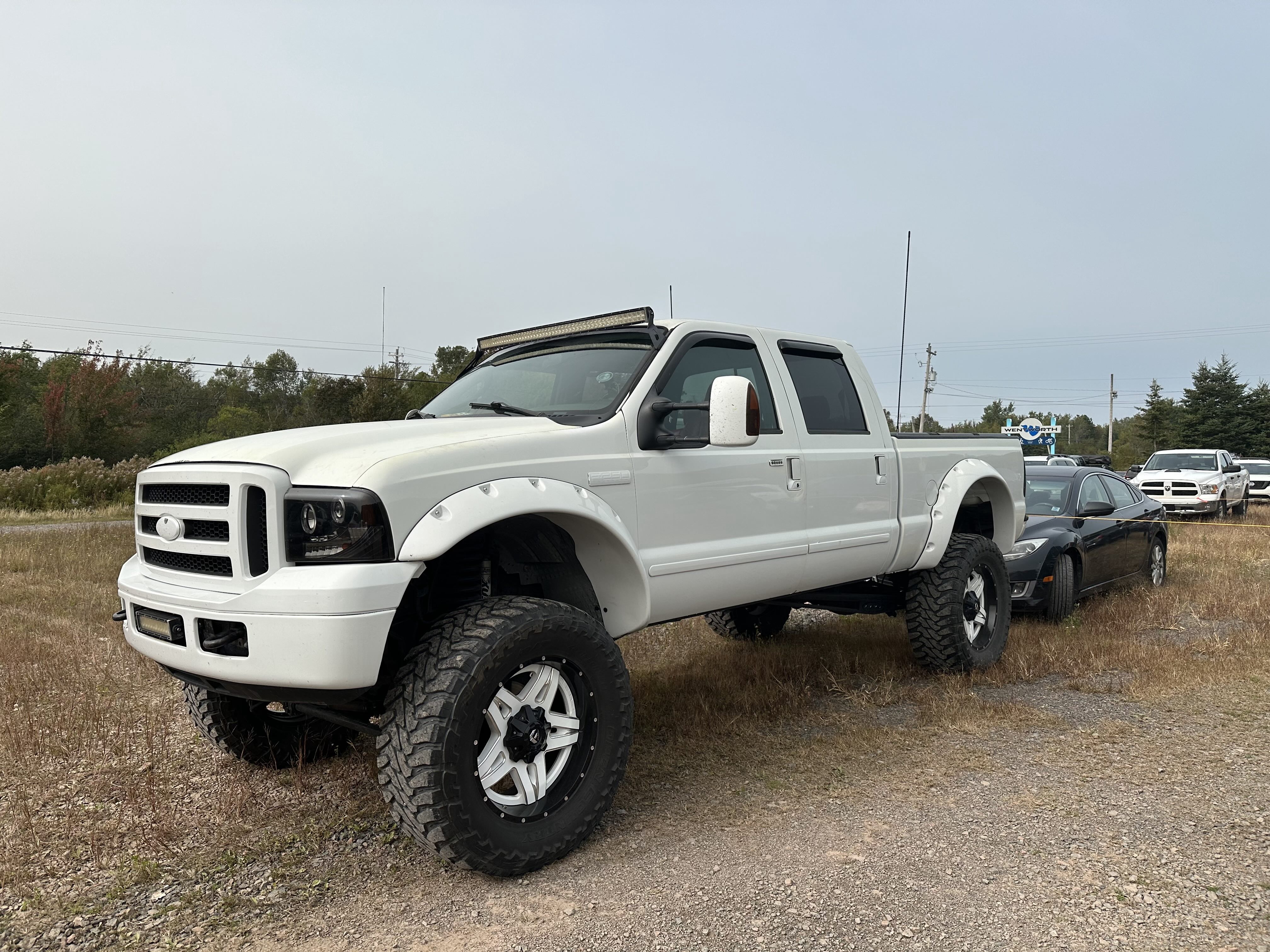 A giant white pickup truck with raised suspension and massive wheels is parked in front of a small black car on a gravel parking lot.