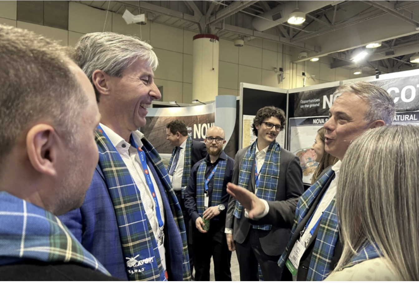 A man with grey hair, a Nova Scotia blue and yellow tartan scarf, and a blue jacket with a white shirt, is shown from the side, and he appears to be laughing loudly, at something several other men (also wearing NS tartan scarfs) are saying to him.