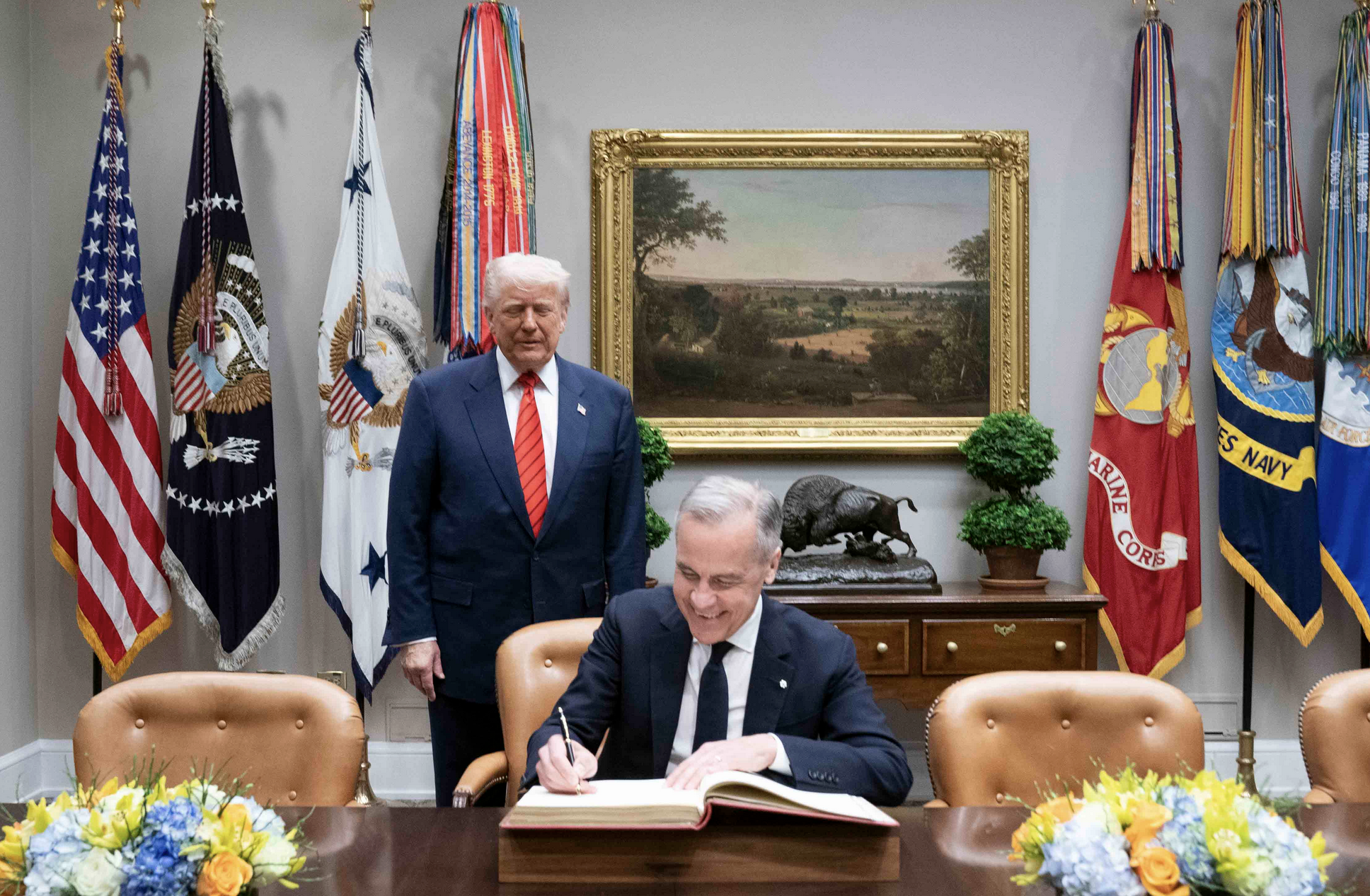 A grey-haired man wearing a navy blue suit and red tie stands unsmiling behind another grey-haired man, with a wide smile, who is seated on a brown upholstery chair and signing a large red book. In the foreground on the desk are bouquets with orange, blue, and yellow flowers. On the wall behind the standing man is a painting of a landscape in a gilt frame, and on either side, an array of drooping flags, including the American stars and stripes on teh far left.